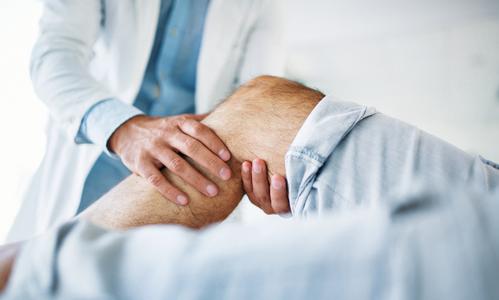 Closeup side view of a doctor examining the knee of a male patient during an appointment. The doctor is gently touching the tendons around the knee and the knee cap and trying to determine the cause of pain.