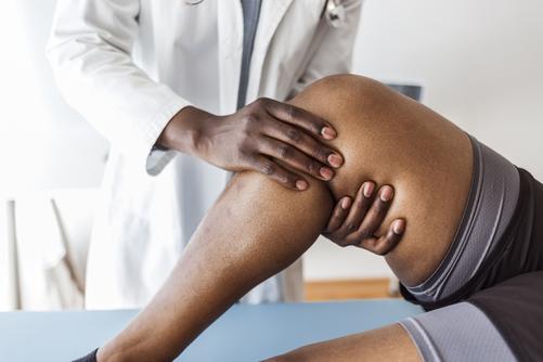 Closeup side view of a doctor examining the knee of a woman during an appointment. The doctor is gently touching the tendons around the knee and the knee cap and trying to determine the cause of pain.
