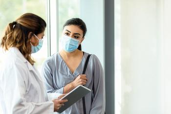 A female doctor explains test results to a middle aged female patient. The results are displayed on a digital tablet. Both women wear masks because of COVID-19.