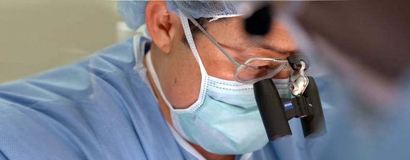 Close up photo of a surgeon working in an operating room while wearing a mask, hair net and magnifying glasses.