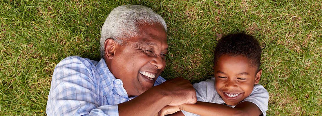 A grandfather tickles his grandson as they lay on the grass outdoors.
