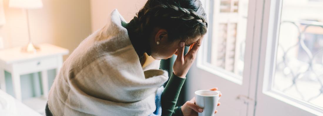 A woman feeling sick is staying home wrapped in a blanket and drinking hot tea.
