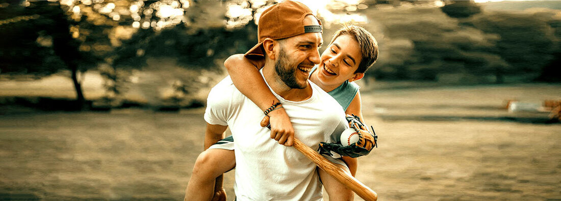 A father carries his son on his back after playing baseball in a park.