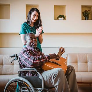 坐在轮椅上的非洲裔老年病人正在给身穿绿色工作服的女医护人员弹吉他。世界杯欧洲区附加赛