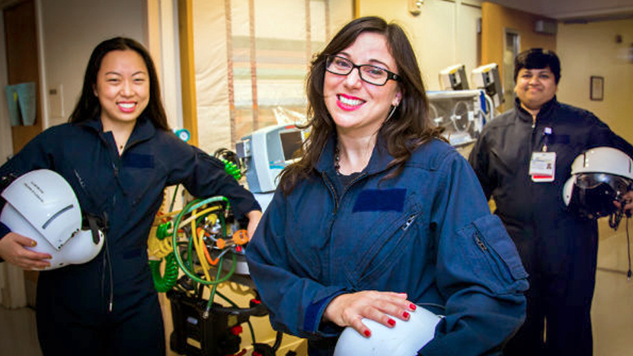 A team of 3 female rapid response physicians stand in the neonative intensive care unit. They are holding their helmets and wearing flight suits.