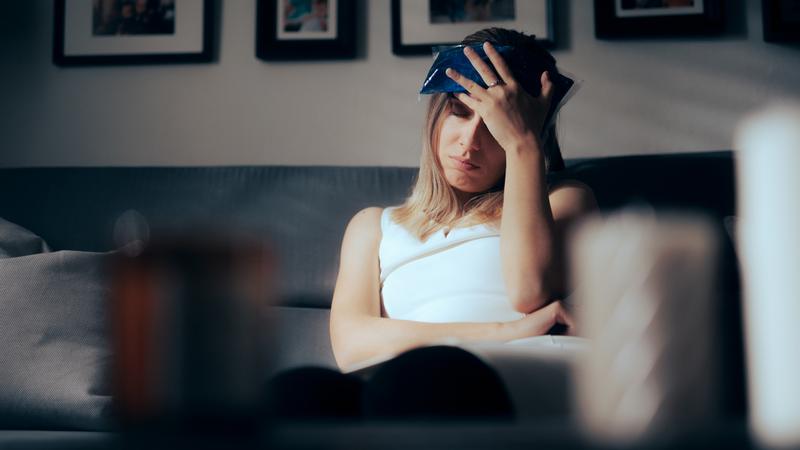 A woman sits on her couch and holds an ice pack on her head while suffering from a migraine.