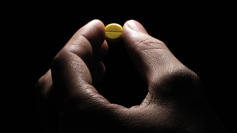 Close up photo of a hand holding a single pill, with dramatic lighting.