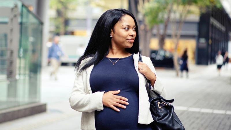 A professional African American woman has her hand on her pregnant belly as she walks in a city.