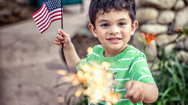 A young boy holds an American flag and a sparkler on the 4th of July.