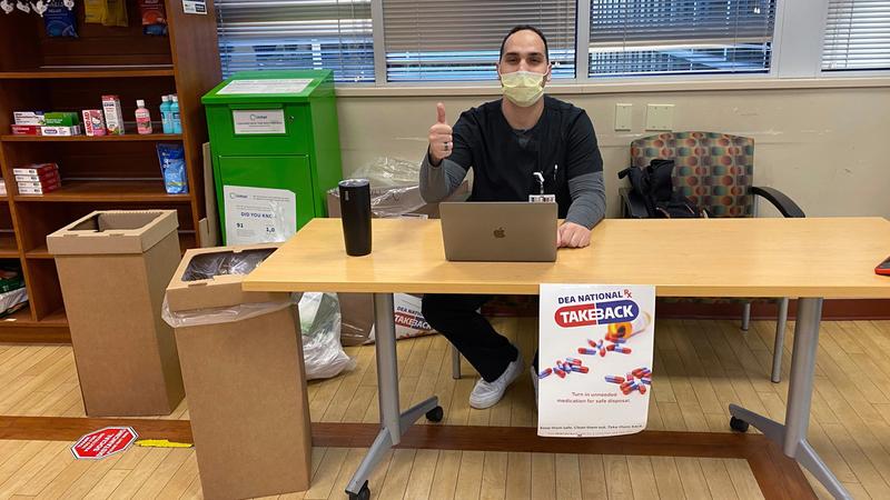 A MedStar pharmacist wearing a mask, sits at a table and gives a thumbs up at an event to take back unused drugs and pharmaceutical paraphenalia for safe disposal.