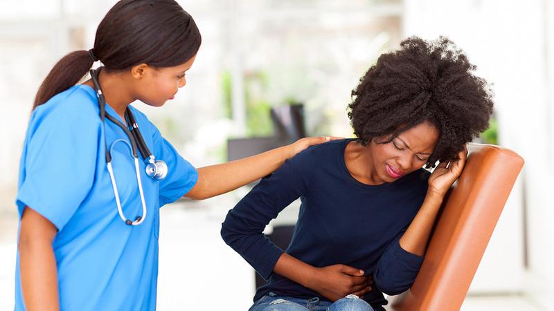 A female patient winces in pain while she holds her abdomen while a healthcare professional leans in to talk to her.