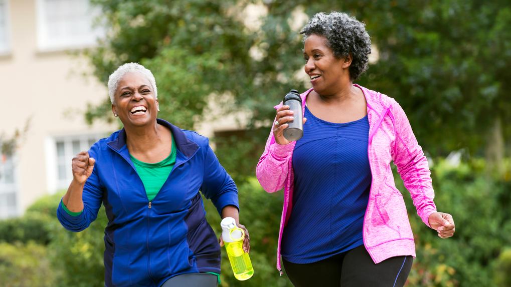 Two senior African American women getting in shape together. They are jogging or power walking on a sidewalk in a residential neighborhood, talking and laughing.