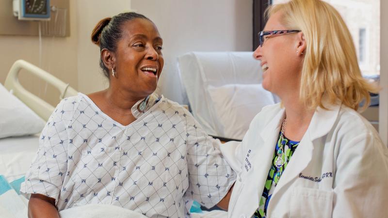 A doctor, wearing a white lab coat, sits on the edge of a patient's bed as they laugh and talk together.