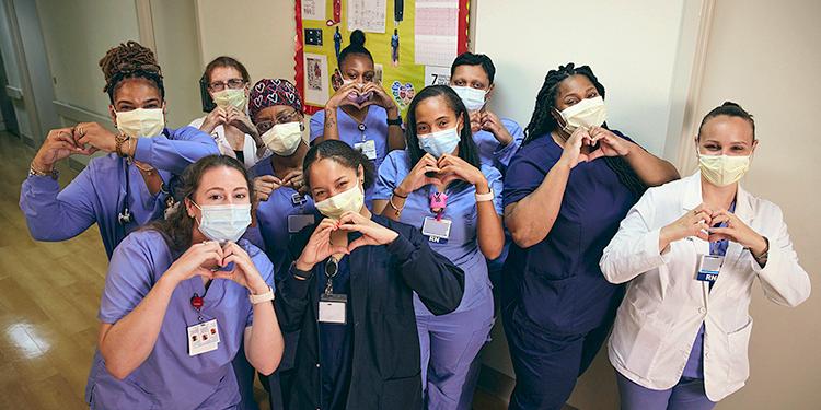 A group of nurses at MedStar Southern Maryland Hospital Center stand together for a group photo and hold their hands in a heart shape. Everyone pictured is wearing a mask.