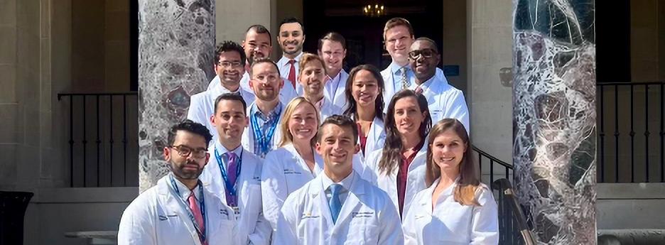 A group of residents wearing white lab coats stand and pose for a photo against a blue wall with the MedStar Orthopedic Institute logo on it.