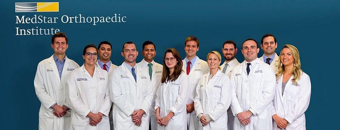 A group of residents wearing white lab coats stand and pose for a photo against a blue wall with the MedStar Orthopedic Institute logo on it.