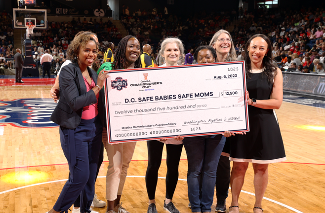 A group of women stand on a basketball court and hold a large donation check for MedStar Health Safe Babies Safe Moms program.