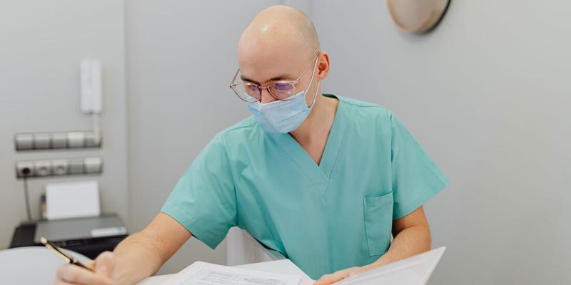A doctor wearing a mask and green scrubs fills out paperwork in an office setting.