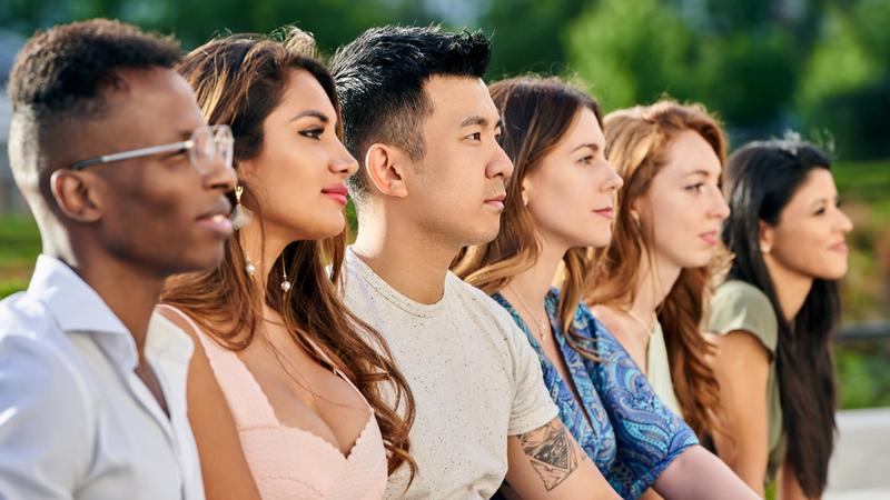 Group of multiracial young people looking away while sitting in row together outdoors.