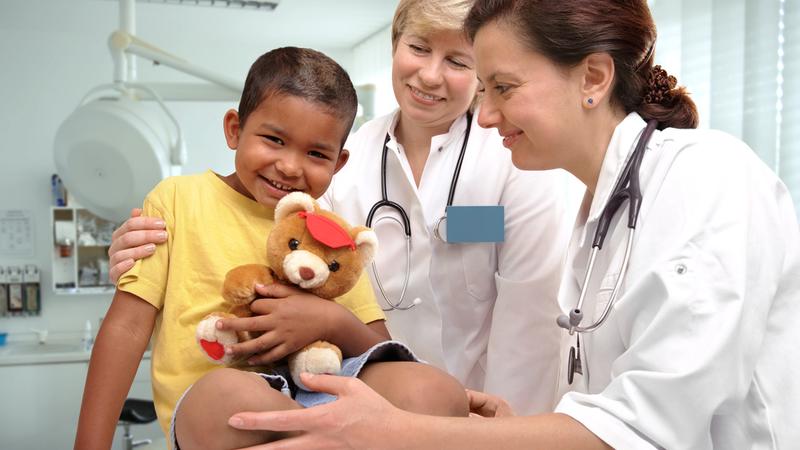 2名女医生在诊所检查一个微笑的孩子，他穿着黄色衬衫，手里拿着泰迪熊。