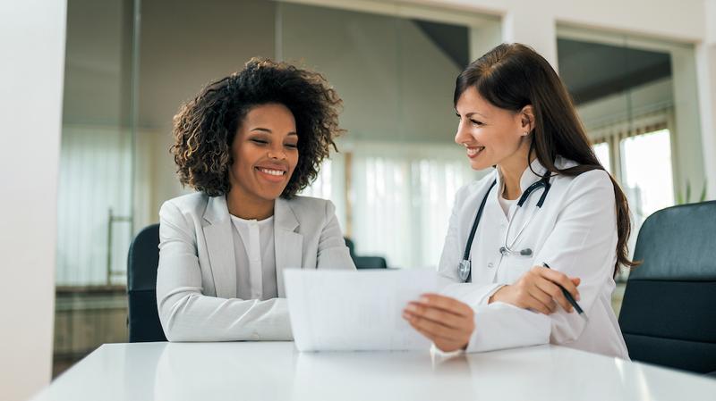 A doctor sits at a table with a patient and shows them an ipad.