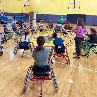 A group of youth wheelchair athletes gather in a gymnasium while a coach speaks to them.