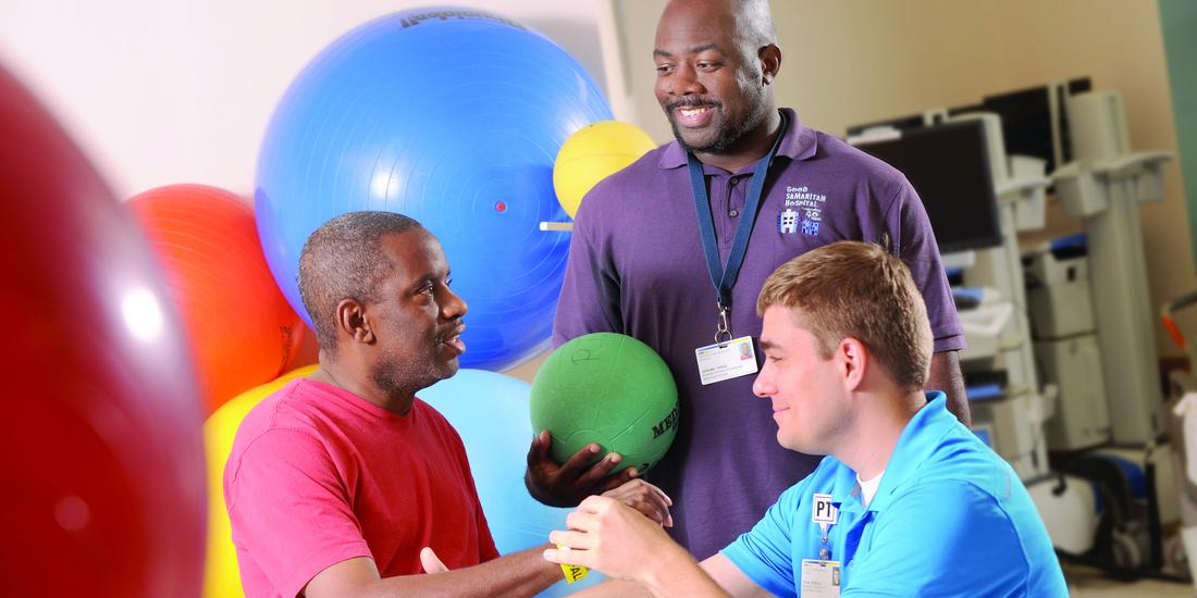 2 physical therapists work with a patient at MedStar Good Samaritan Hospital's rehabilitation center. The patient is seated and there are colorful therapy balls in the background.