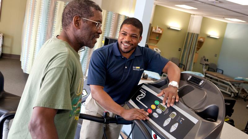 A male patient walks on a treadmill at a MedStar Health rehabilitation gym, while his physical therapist adjusts the controls.