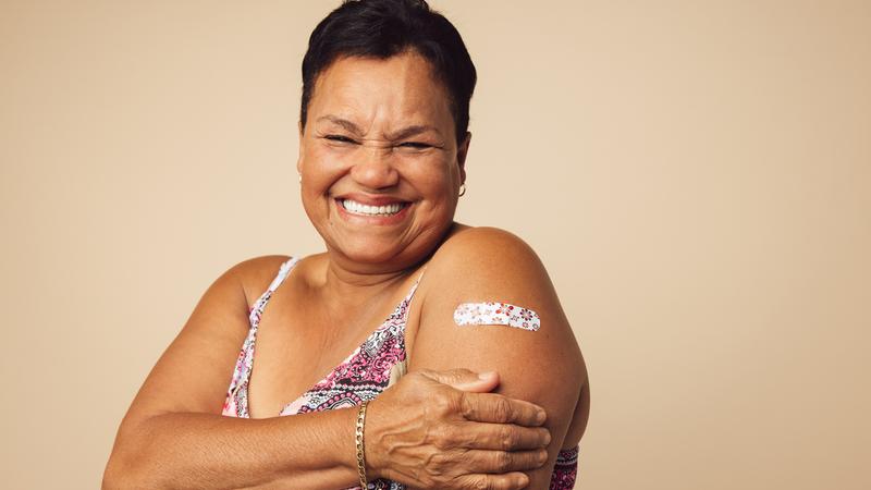 A middle-aged African American woman stands with her shoulder to the camera to show a band-aid on her arm, after receiving a vaccination.