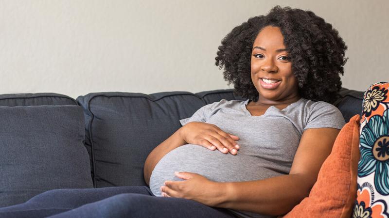 A pregnant woman relaxes on a sofa with her hands on her belly, and smiles for the camera.