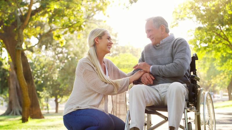 A young caregiver crouches to talk with an older man who is sitting in a wheelchair. They are outdoors in a park.