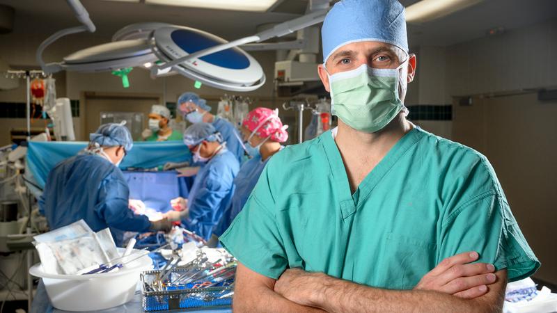 Dr. Christian Schults stands in an operating room and poses with his arms crossed while looking at the camera. He is wearing a green scrubs, a blue mask and surgical skull cap. A surgical team works in the background.