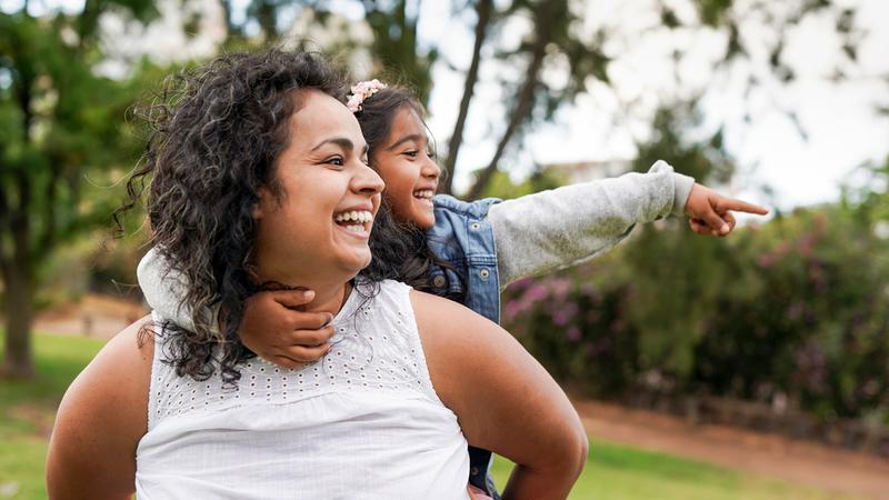 A woman holds a young child on her back. The child is pointing at something to the side and they are both smiling.