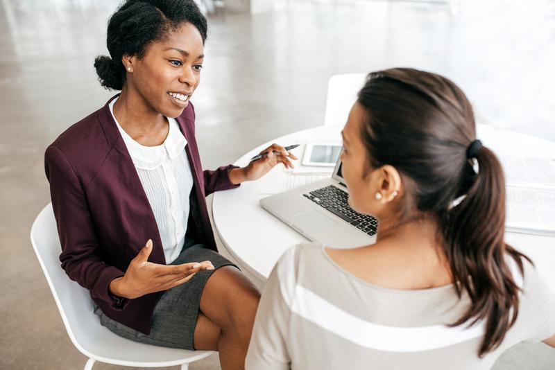 Two professional women sit together at a table and have a discussion in an office setting.