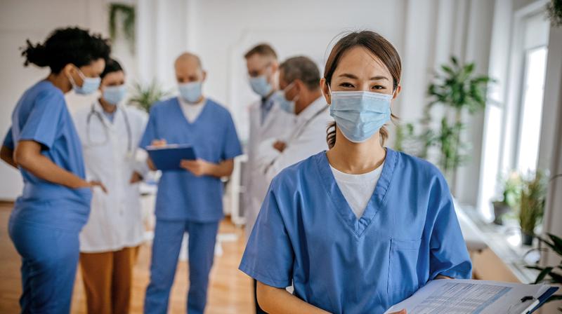 A nurse wearing a face mask stands and looks at the camera, while a group of healthcare professionals is out of focus in the background.