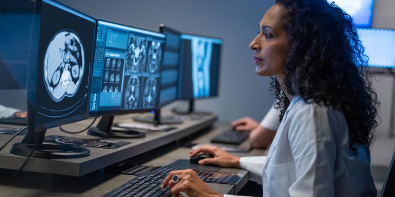 A female physician looks at a row of computer screens showing diagnostic radiology scans.