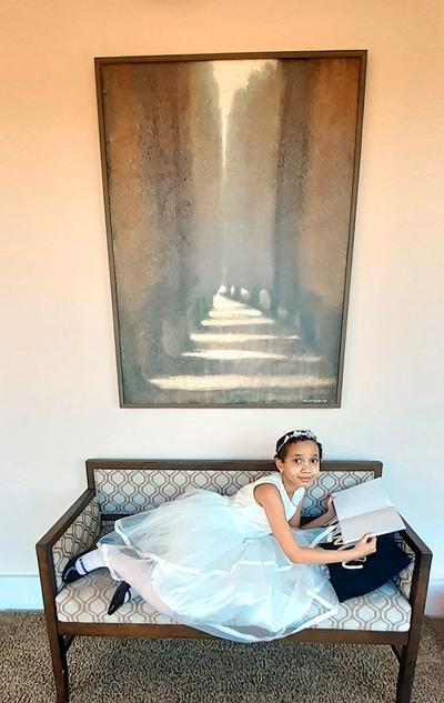 A little girl in a fancy white dress lays on a bench beneath a painting and reads a book.