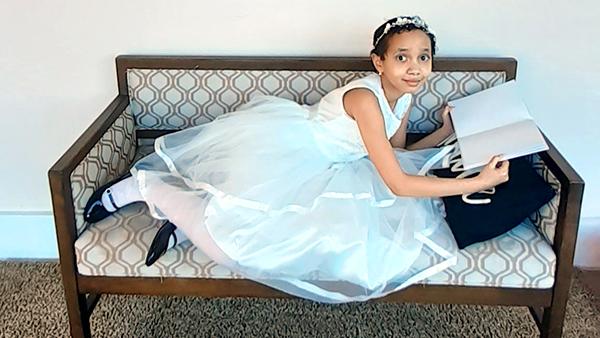 A little girl in a fancy white dress lays on a bench beneath a painting and reads a book.