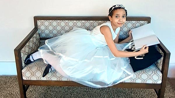 A little girl in a fancy white dress lays on a bench beneath a painting and reads a book.