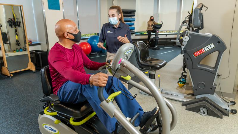 A female physical therapist works with a male patient who is riding a stationary bike in a rehabilitation gym.