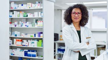 A female pharmacist stands in a pharmacy looking at the camera and smiling with her arms crossed.