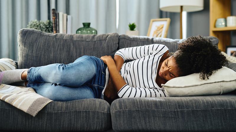 A woman lays on a sofa and holds her stomach as if in pain.