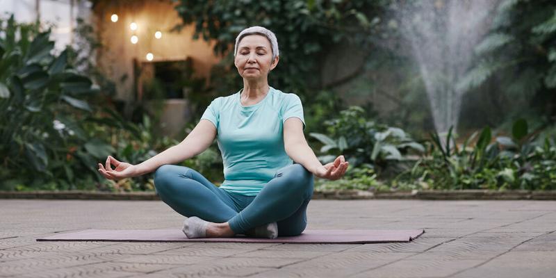 A senior woman sits in a meditative yoga position in a garden outdoors.