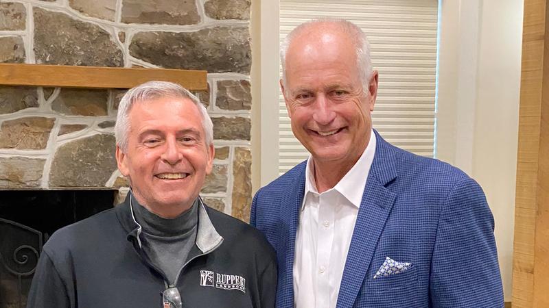 Craig Ruppert and Bill Law stand side-by-side and pose for the camera. They are indoors standing in front of a stone fireplace.