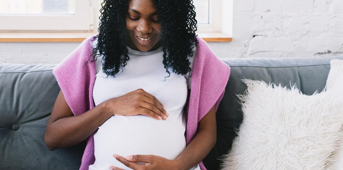 An expectant mother sits on a couch and looks down at her hands on her pregnant belly