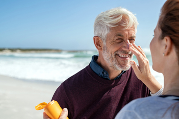 Portrait of cheerful older man looking at his mature wife applying sunscreen on nose. Senior husband enjoying vacation with woman while applying sunscreen on face at beach. Middle aged retired couple applying suntan lotion at sea.