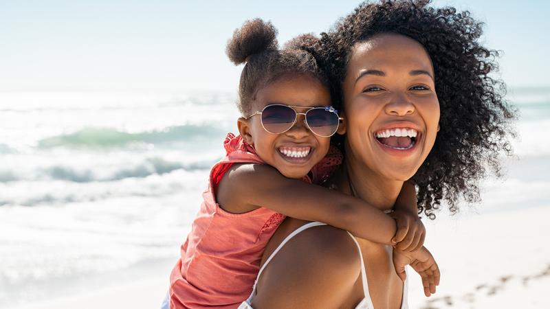 一个年轻的女人拥有一个小女孩在她的背上,因为他们在海滩上玩得开心。他们笑眯眯地看着镜头,。