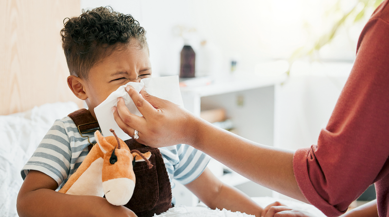 A mother holds a tissue to her young son's nose so he can blow his nose.