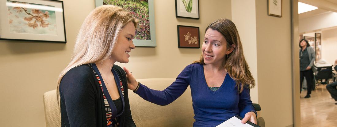 A female doctor rests her hand on her female patient's shoulder as they sit together and talk in the office waiting room. 