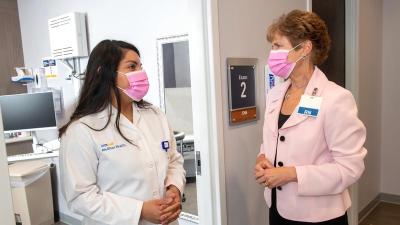 Jennifer Daniella Son and Cathy Brophy, both wearing masks, stand and talk in the hallway outside of a patient exam room.